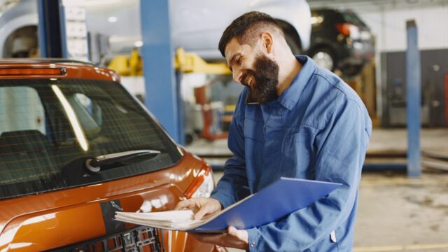 Smiling Man in Blue Coveralls Standing Beside an Orange Car Having Conversation Over the Phone